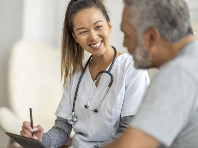 young nurse smiling at older man as he talks to her, she is taking notes on a clipboard